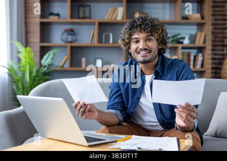 A smiling man is looking at the camera holding papers in front of a laptop while working remotely from home. Stock Photo