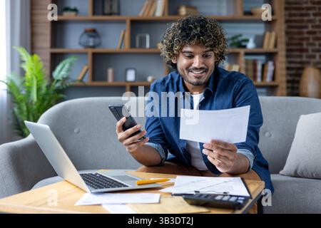 A happy man examines a bill while using his smartphone and laptop, working on his finances from home. Stock Photo