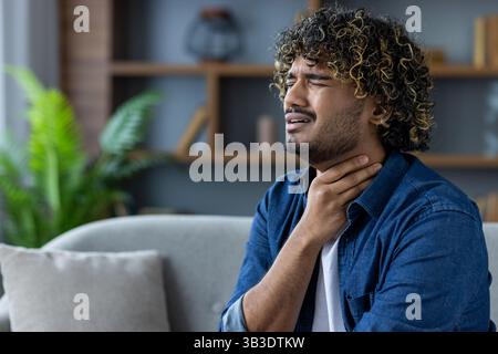 A man with curly hair holds his hand to his throat, seemingly in pain while sitting in a living room. Stock Photo