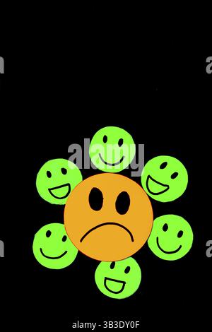 Vertical image of stickers or paper cut of a sad yellow face surrounded by smiling and happy green faces on a black background with copy space Stock Photo