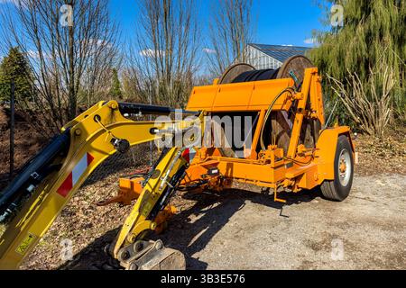 A yellow excavator stands next to a large orange machine with wooden cable reel. The scene reflects a sunny day, with clear skies and trees in the bac Stock Photo