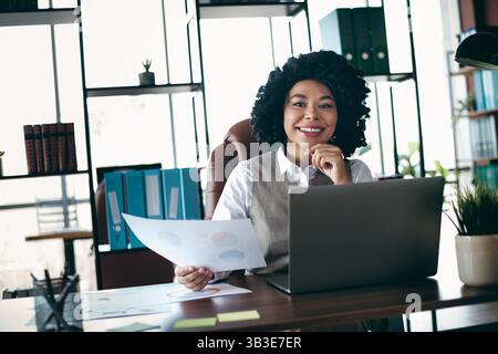 Skilled businesswoman in a professional office working on a laptop while reviewing documents and smiling confidently Stock Photo