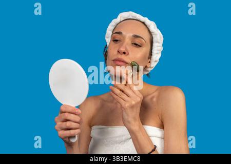Woman wearing bathrobe and hair band using jade roller massaging her face looking in a mirror on a blue background Stock Photo