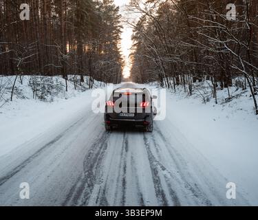 Peugeot 5008 driving into sunset in snowy woodland. Rear end view of dark grey car driving on a narrow road through winter forest, surrounded by pine Stock Photo