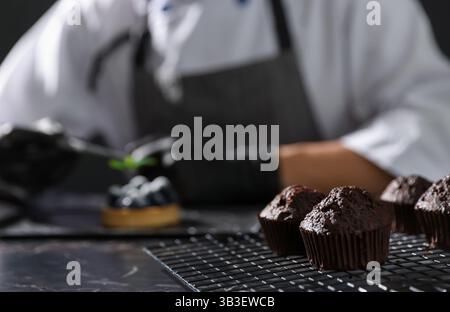 Confectioner decorating tasty tart with mint at dark table, focus on chocolate muffins Stock Photo