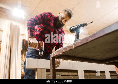 Skilled carpenter constructs wooden furniture in workshop using power tools during daytime hours Stock Photo