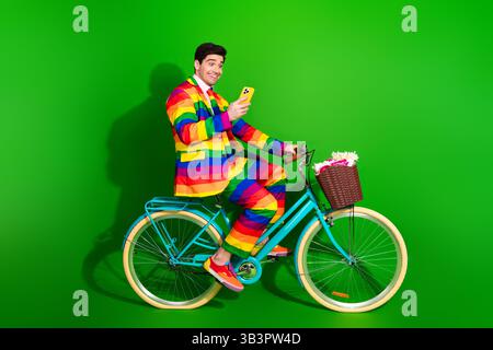 Young man in vibrant rainbow suit on bicycle with green backdrop portraying pride, happiness, and fashionable style Stock Photo