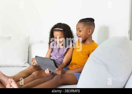 Smiling Diverse siblings using tablet device on light-colored sofa in living room, with cushions Stock Photo