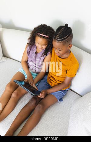 Diverse siblings interacting with black tablet while sitting on light sofa, exploring content Stock Photo
