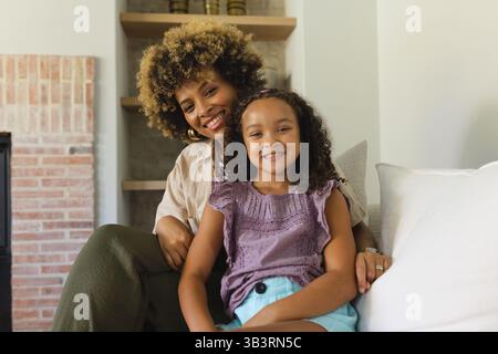 Smiling Diverse mother and daughter sitting on couch in living room, with decorative shelving unit Stock Photo
