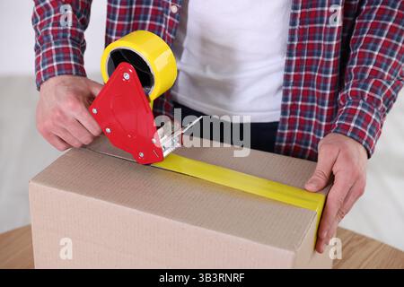 Man using dispenser with adhesive tape to seal cardboard box indoors, closeup Stock Photo