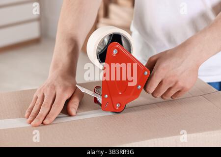 Man using dispenser with adhesive tape to seal cardboard box indoors, closeup Stock Photo