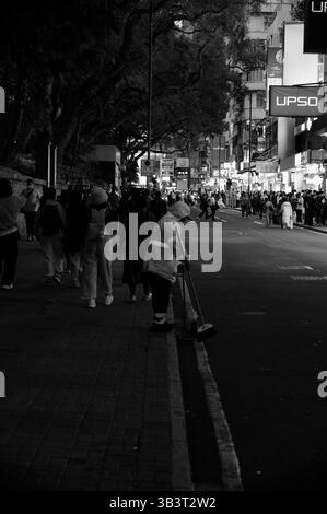 Street cleaner in reflective gear quietly tends the curb as crowds move through a busy night Stock Photo