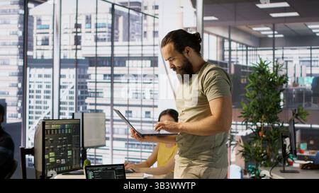 Admin at office desk utilizing cloud computing resources on laptop to train large scale machine learning models. Man monitoring company systems using notebook while standing in workspace, camera A Stock Photo