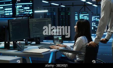 Person with paraplegia helped by coworker in data center to arrive at desk, coding on PC. Woman in wheelchair doing maintenance on server room infrastructure rackmounts, camera A Stock Photo