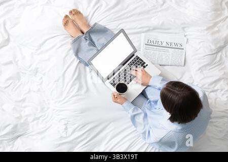Woman in pajamas using laptop with coffee cup and newspaper on bed, top view Stock Photo