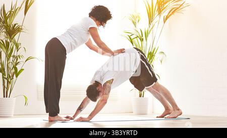Female yoga instructor training a man to do downward dog pose during a fitness class in an exercise studio. Yogi helping to correct body form and Stock Photo