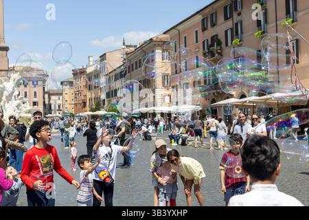Rom, Italien - April, 23, 2025: In a bustling square, children run and laugh as they chase colorful bubbles floating in the air. The warm sun shines d Stock Photo