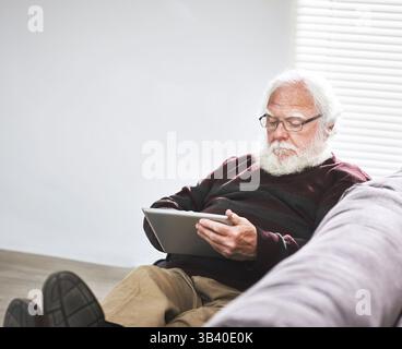 A relaxed senior man typing on a tablet and relaxing on the floor at home enjoying retirement. A mature man browsing the internet learning to use Stock Photo
