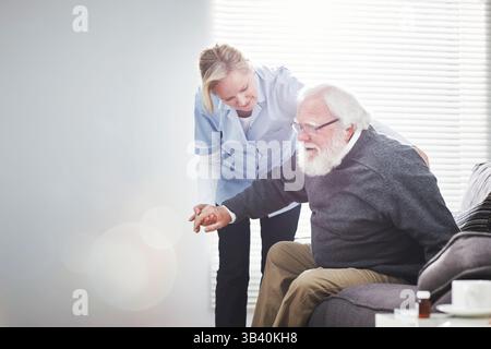 Medical doctor, nurse or female worker helping a senior man to sit, relax or get rest on the couch at home. Sick, frail and retired male getting help Stock Photo