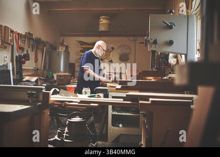 Old carpenter busy working in his workshop on a project or design. Senior man enjoys physical labour and using his hands to make furniture Stock Photo