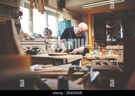 A senior handyman working in his home garage or workshop cutting or sanding wood. Skilled mature male craftsman building, repairing or cutting a piece Stock Photo