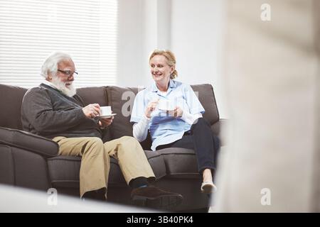 Relaxed senior interracial couple having tea and talking about old happy memories while relaxing on a couch at home. A mature man and woman drinking Stock Photo