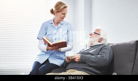 Nurse or caregiver reading a book or bible to her senior patient having pleasant and warm conversation on a couch. Female heathlcare aid bonding and Stock Photo
