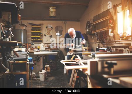Working mature senior carpenter building, planning and making furniture indoors in a workshop. Elderly man work, make and design a wood chair. Old Stock Photo