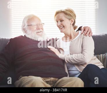 Affectionate senior couple relaxing, bonding and laughing in their living room at home. Mature, romantic and loving husband and wife cuddling and Stock Photo