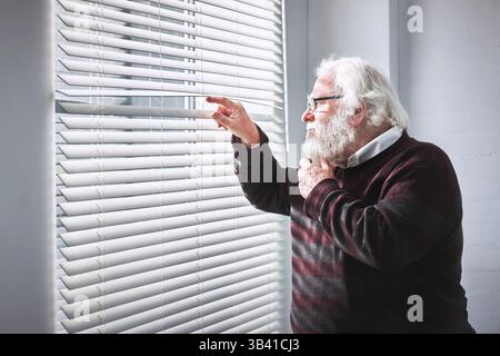 Curious senior man looking through window blinds while thinking, planning and wishing to escape old age home. Stressed, anxious or lonely elderly Stock Photo