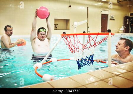 Disability, fitness and swimming activity by down syndrome man playing basketball in indoor pool. Care facility for special needs people training in Stock Photo