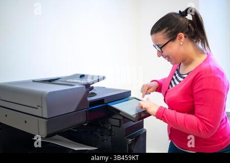 Smiling office worker interacting with the touchscreen of a modern multifunction printer, configuring a print job and checking the device's status for Stock Photo