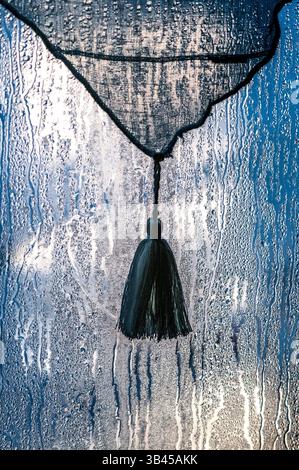 Tassel on net curtain behind rain-splashed window in morning light. Stock Photo