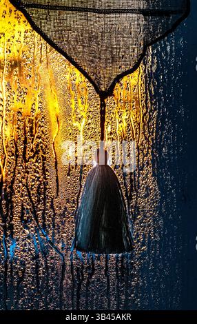 Tassel on net curtain behind rain-splashed window in evening light. Stock Photo