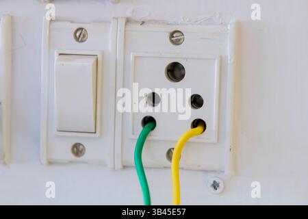 Colorful green and yellow wires connect to an electrical socket near a light switch on a white wall. Stock Photo
