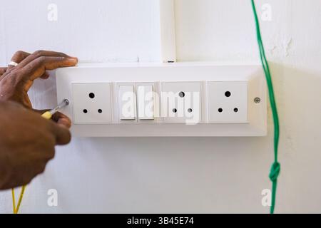 A skilled electrician works on installing electrical outlets and switches on a white wall indoors during daylight hours. Stock Photo