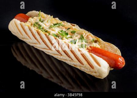 Grilled hot dog in toasted bun with fresh vegetable salad topping, isolated on black background, close-up view. Stock Photo