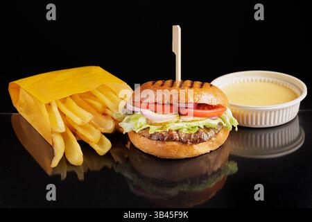 Delicious cheeseburger with fresh fries in yellow paper bag and dipping sauce on black reflective background. Stock Photo