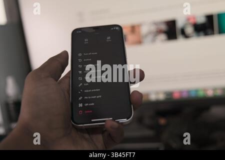 A hand holds a smartphone displaying app features while a computer screen is visible in the background. Stock Photo