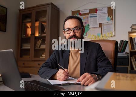 Confident mature school psychologist in eyeglasses looking at camera while sitting by his workplace and taking notes Stock Photo