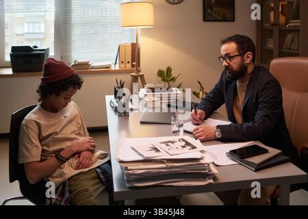 Bearded psychologist taking notes and looking at upset schoolboy sitting in front of him during their conversation at session Stock Photo