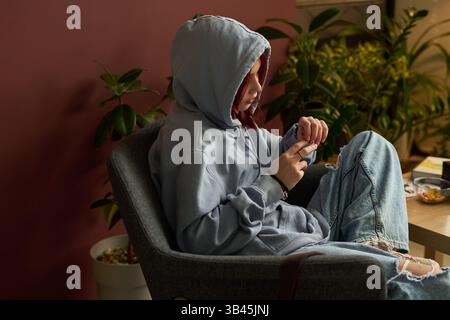 Girl in blue jeans and hoodie talking to school psychologist during session while sitting in armchair against green plants Stock Photo