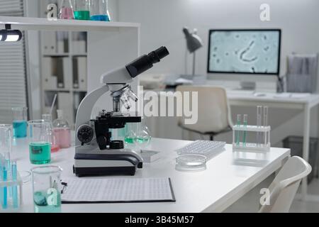 Modern laboratory setup featuring high-tech microscope and various chemical solutions on workbench. Background includes organized workspace with computer showing magnified cellular image Stock Photo