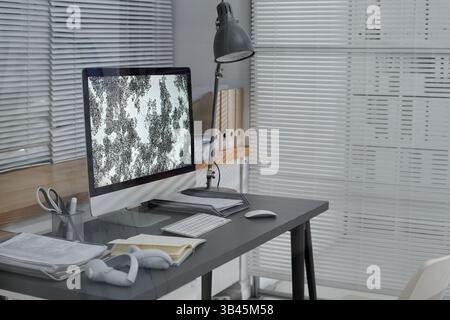 Modern workspace featuring a desktop computer with intricate design display, headphones, office supplies, and a lamp on a sleek desk. White blinds cover large window in background Stock Photo