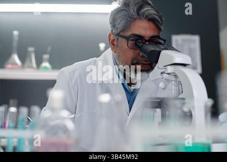 Middle-aged scientist examining samples under microscope in laboratory focusing on research. Various lab equipment and test tubes visible in background Stock Photo