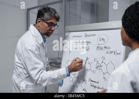 Scientist explaining complex chemical reactions on whiteboard while colleague taking notes focusing on detailed formulas and molecular structures in laboratory setting Stock Photo