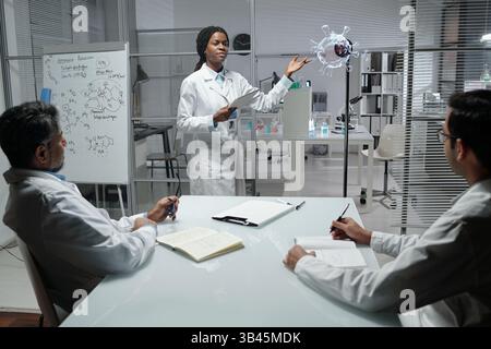 Scientist explaining complex molecular structures during presentation in laboratory setting with colleagues observing and taking notes Stock Photo