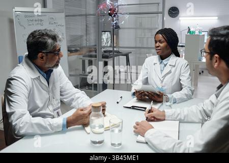 Medical professionals collaborating during a meeting around table in a modern office. Discussion involving diverse team members with tablets and notes on whiteboard in background Stock Photo