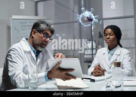 Two scientists in lab coats sitting at a table and discussing research findings on a tablet. One scientist pointing at screen while other listens attentively Stock Photo